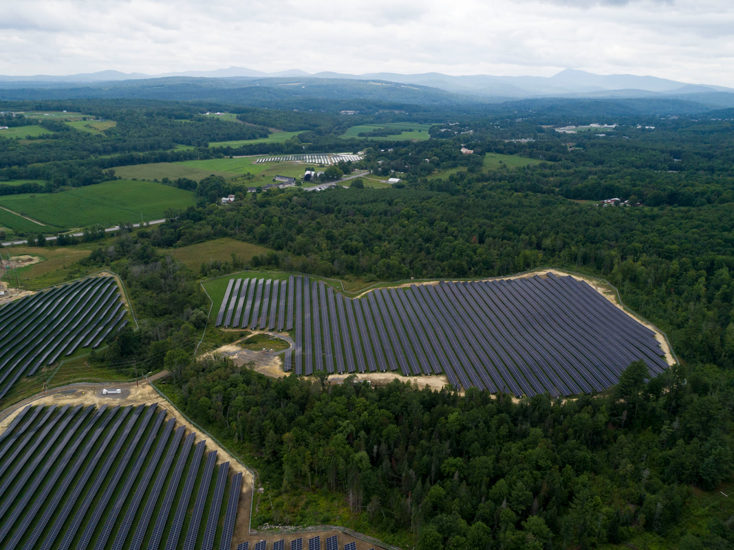 An aerial image of three solar fields surrounded by green forests, with several other green farm plots further back.
