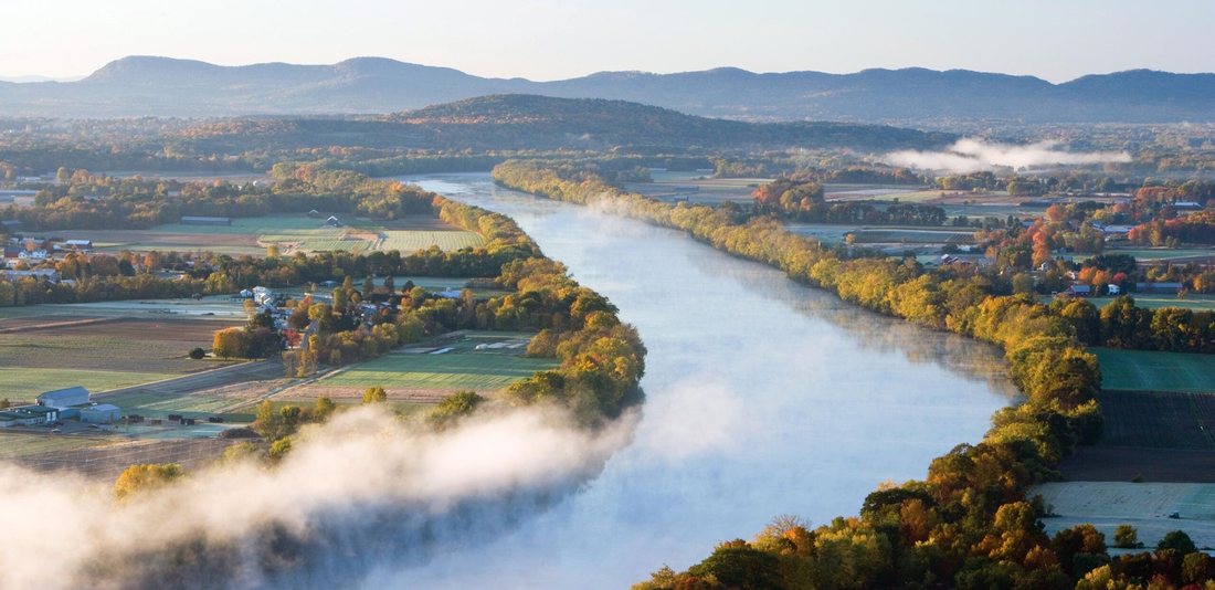 An image of a the Connecticut River, a large, curving body of water lined with autumn trees, various farming plots and fields on either side of the river.